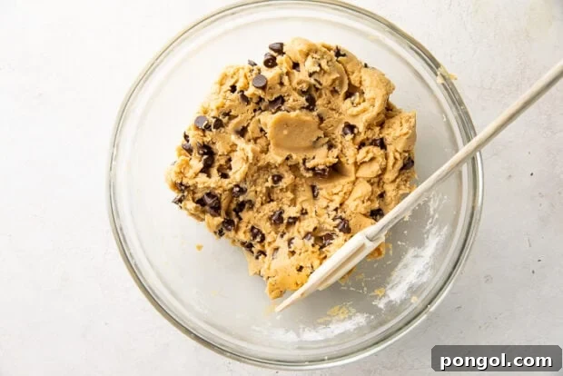 Ultimate Vegan Chocolate Chip Cookies 8 Vegan chocolate chips being stirred into cookie dough in a glass mixing bowl with a spatula.