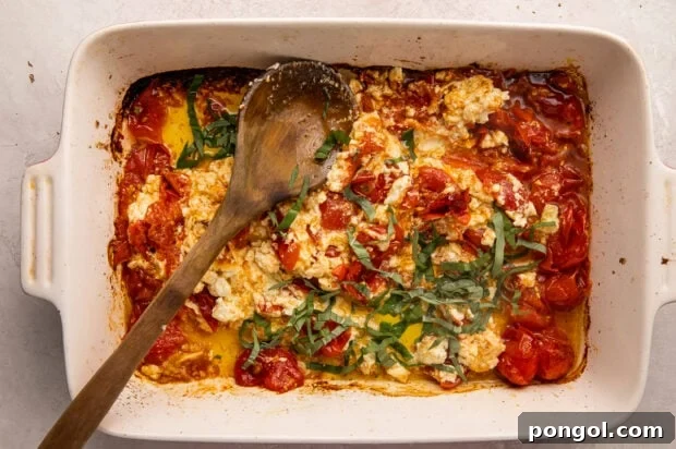 Baked feta and tomatoes, mashed together, with fresh basil and olive oil in a casserole dish, with a wooden spoon laid across diagonally.