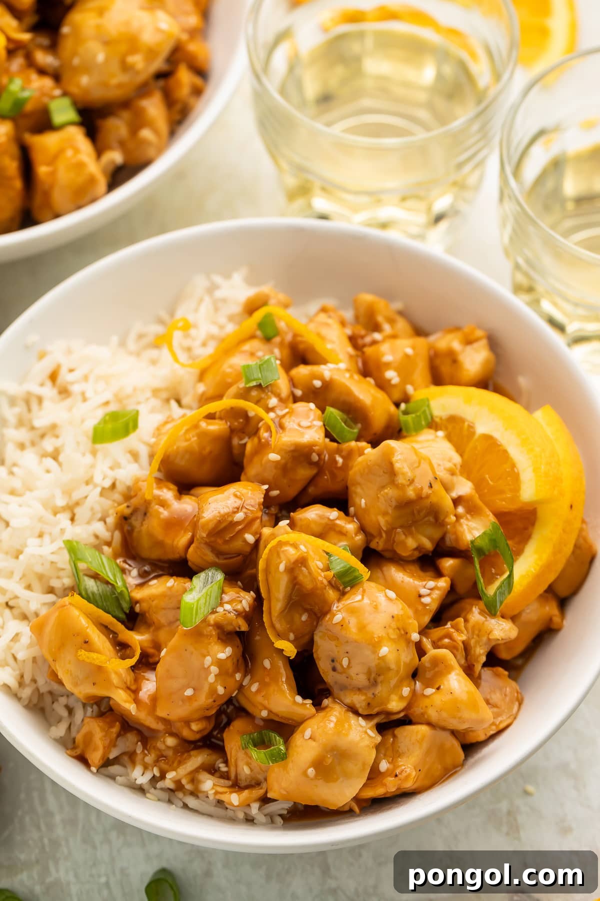 A white bowl filled with white rice, glazed orange chicken, wedges of oranges, and sliced green onions, on a table with glasses of water.