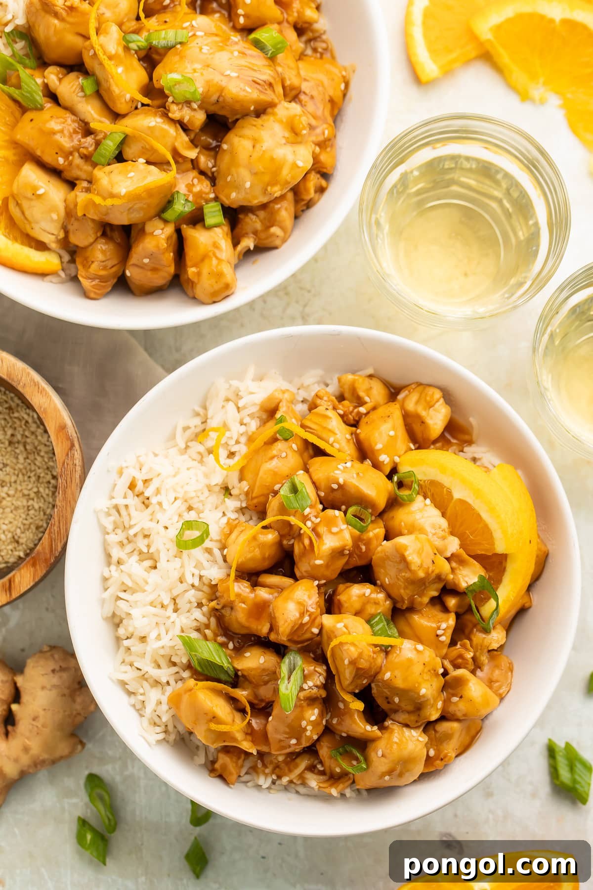 Overhead view of 2 bowls holding fluffy white rice, pieces of glazed orange chicken, orange wedges, and thinly sliced green onions.