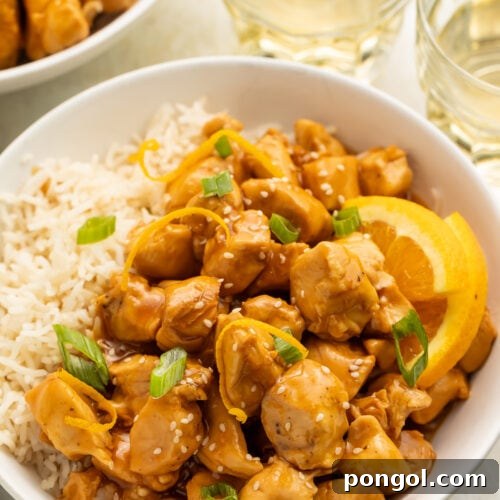 A white bowl filled with white rice, glazed orange chicken, wedges of oranges, and sliced green onions, on a table with glasses of water.