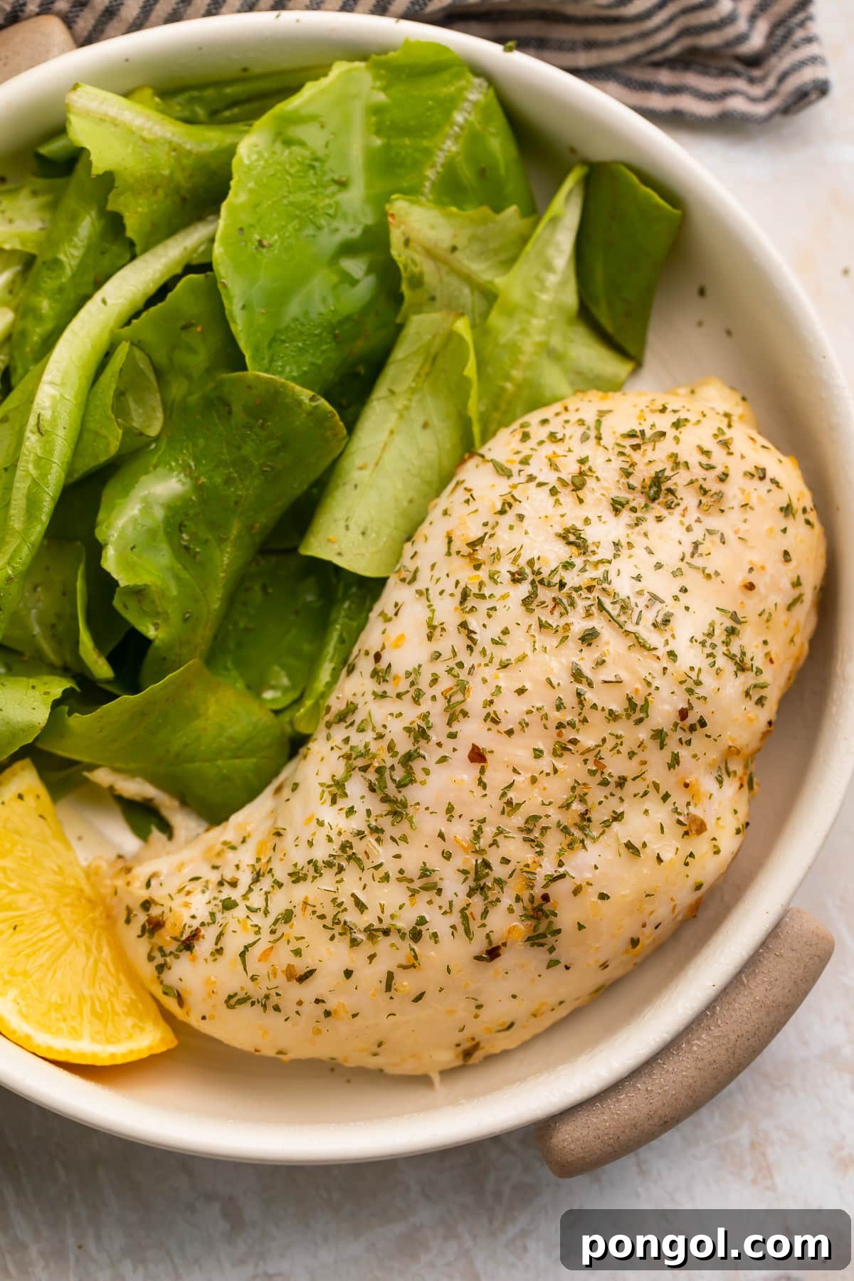 An Italian dressing chicken breast in a shallow bowl with a spinach side salad and a lemon wedge.