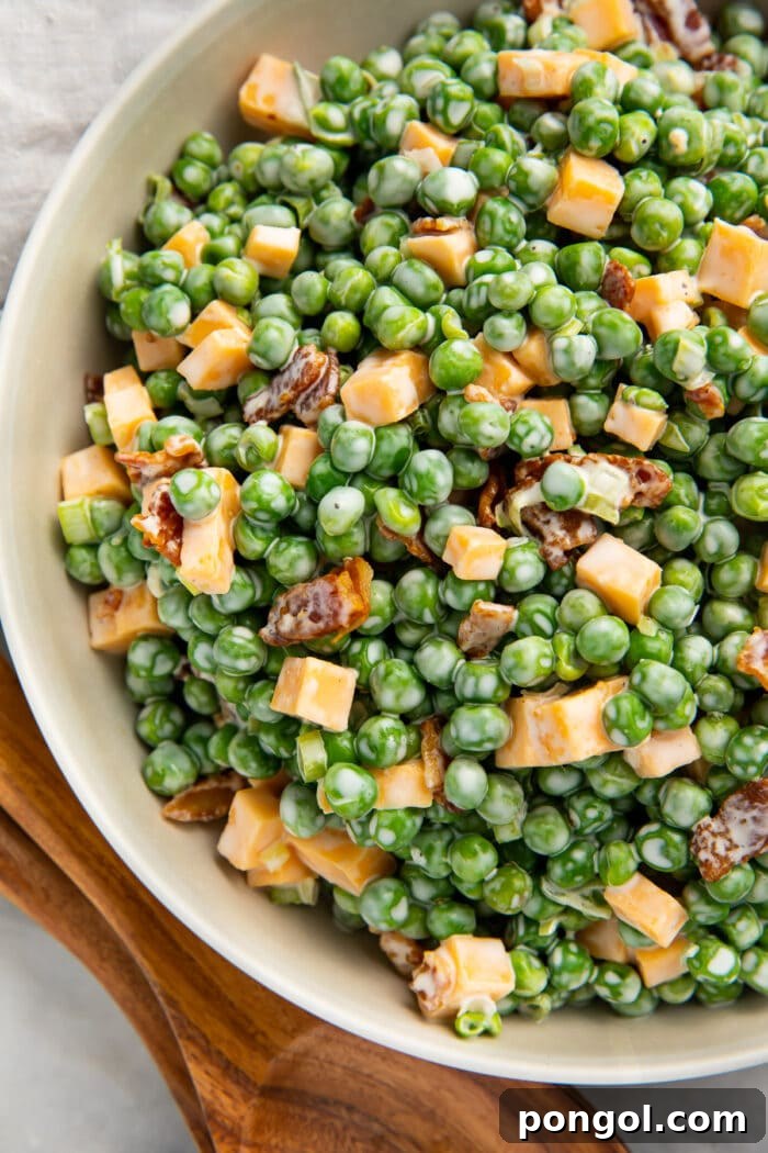 Overhead photo of pea salad with bacon, cheddar cheese, and green onion in a large bowl next to wooden utensils