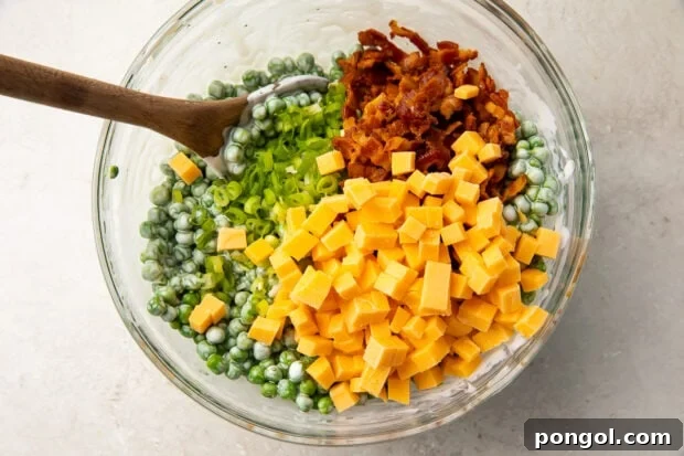 Peas, cheddar, scallions, and bacon in a large glass mixing bowl with a wooden spoon, ready to be mixed.