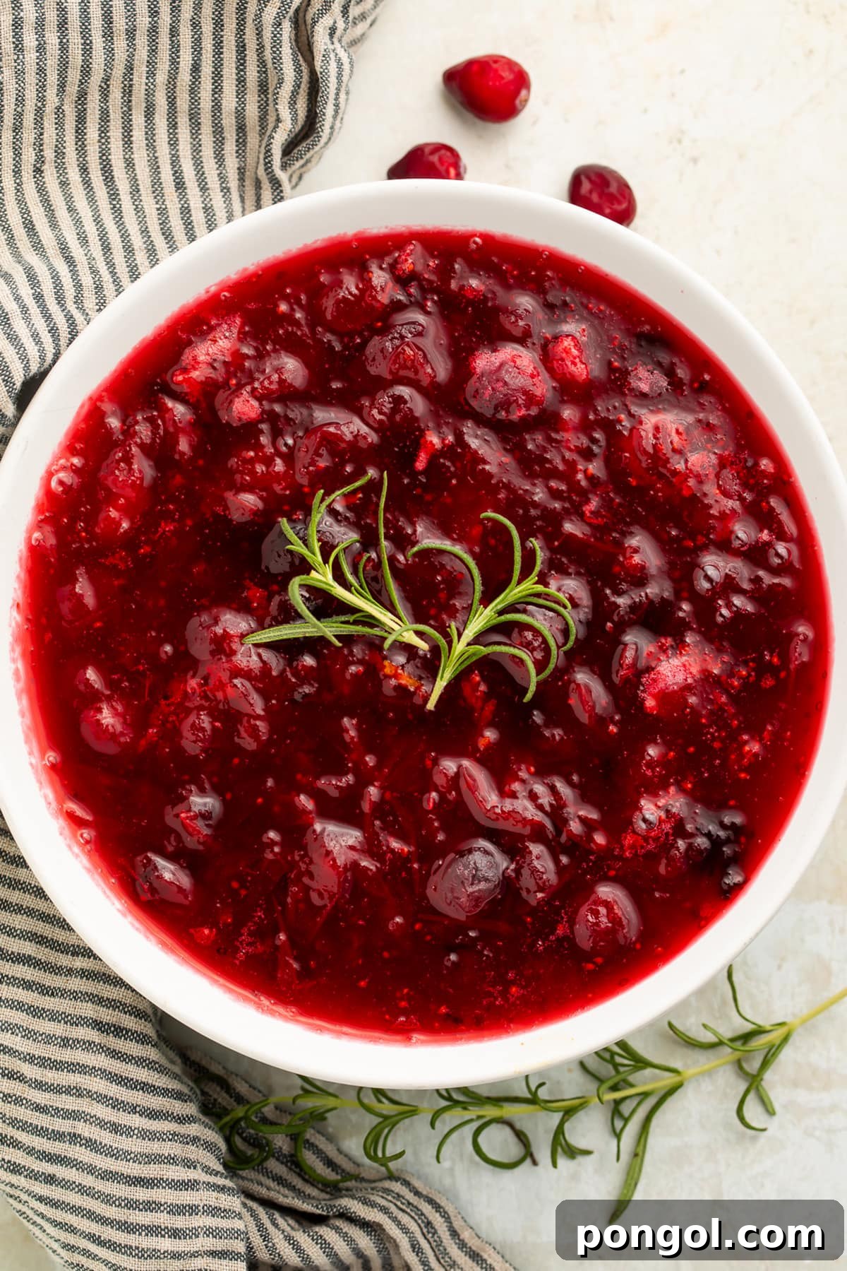Overhead photo of gourmet whole berry rosemary cranberry sauce in a white serving bowl, adorned with a fresh rosemary sprig, showcasing its vibrant color and homemade appeal.