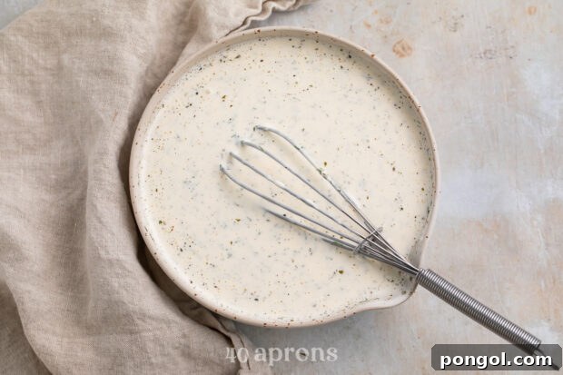 Overhead view of a bowl of buttermilk ranch dressing in a medium mixing bowl with a whisk on a neutral cloth napkin.