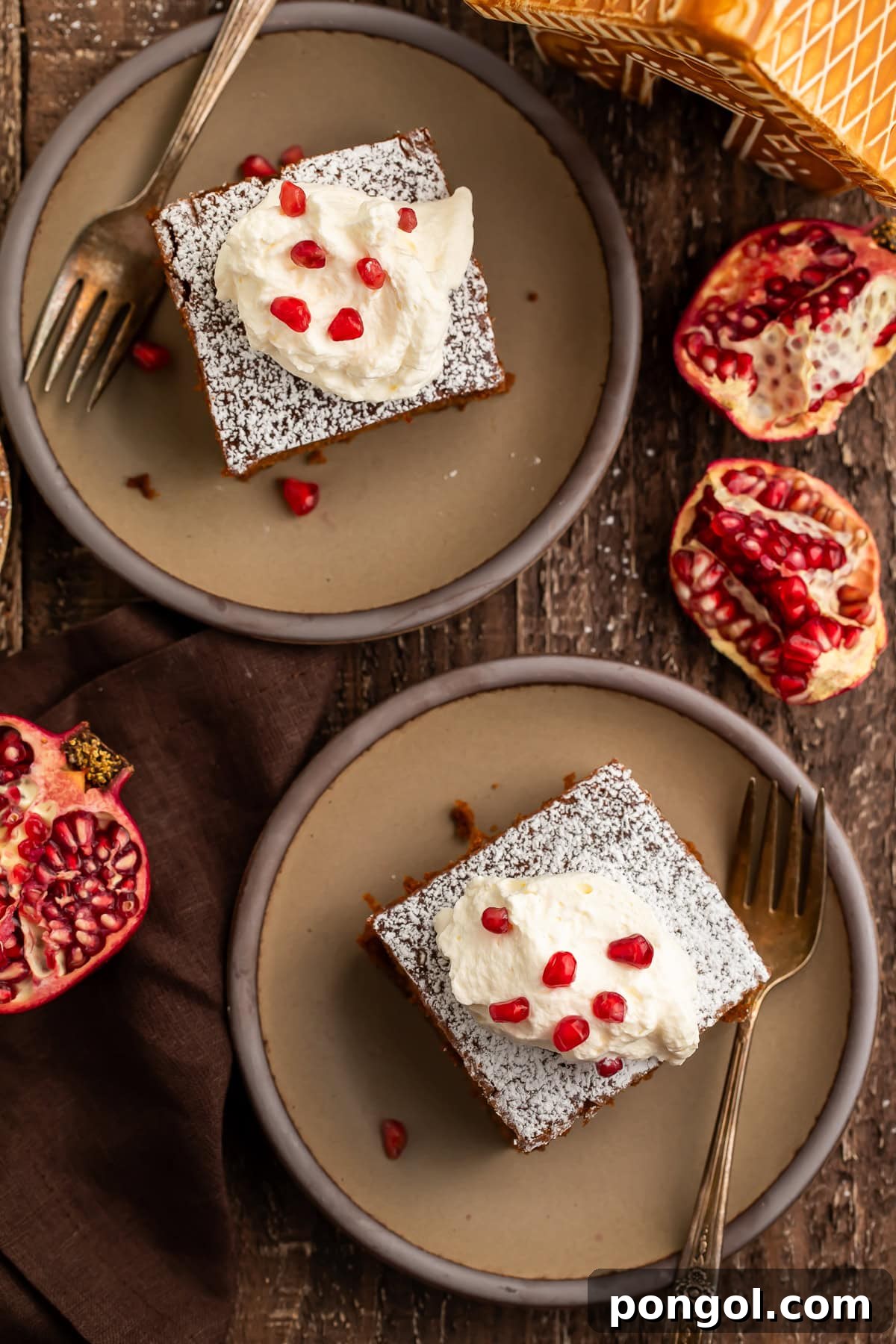Overhead view of two gingerbread cake slices with whipped cream and pomegranate seeds on gray plates.