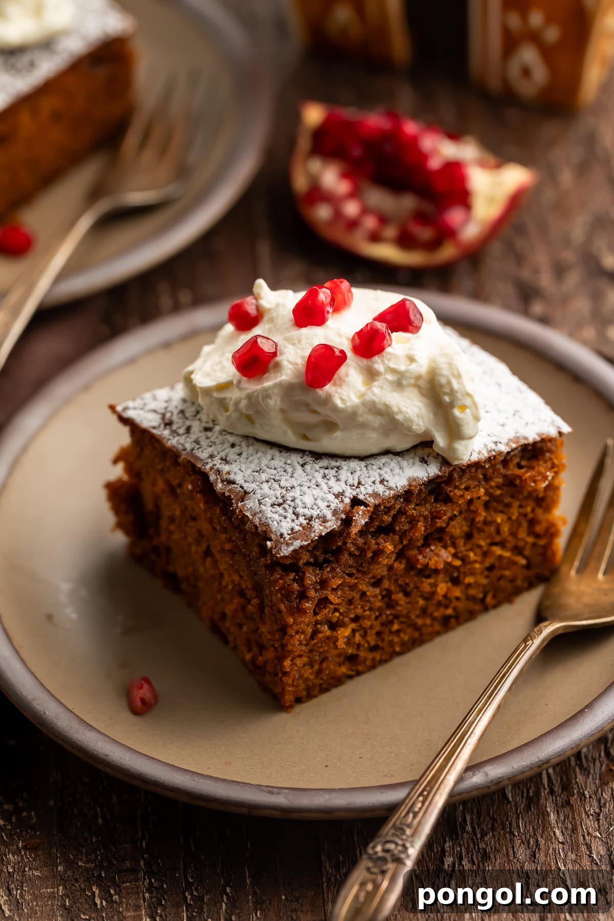 Square gingerbread cake slice with whipped cream dollop and pomegranate seeds on beige plate with powdered sugar.