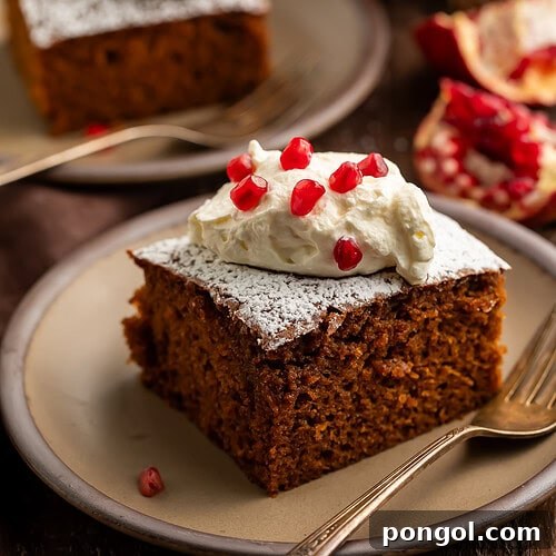 Square slice of gingerbread cake with whipped cream and pomegranate seeds on a beige plate with gold fork.