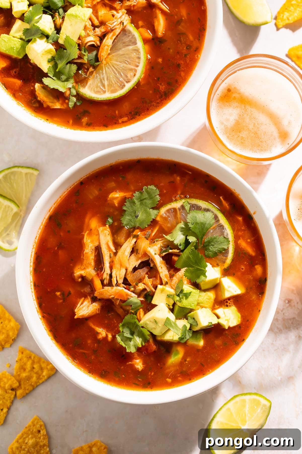 Mexican Chicken Extravaganza 12 Two bowls of Mexican chicken soup on a table, surrounded by lime wedges, tortillas, and glasses of beer.