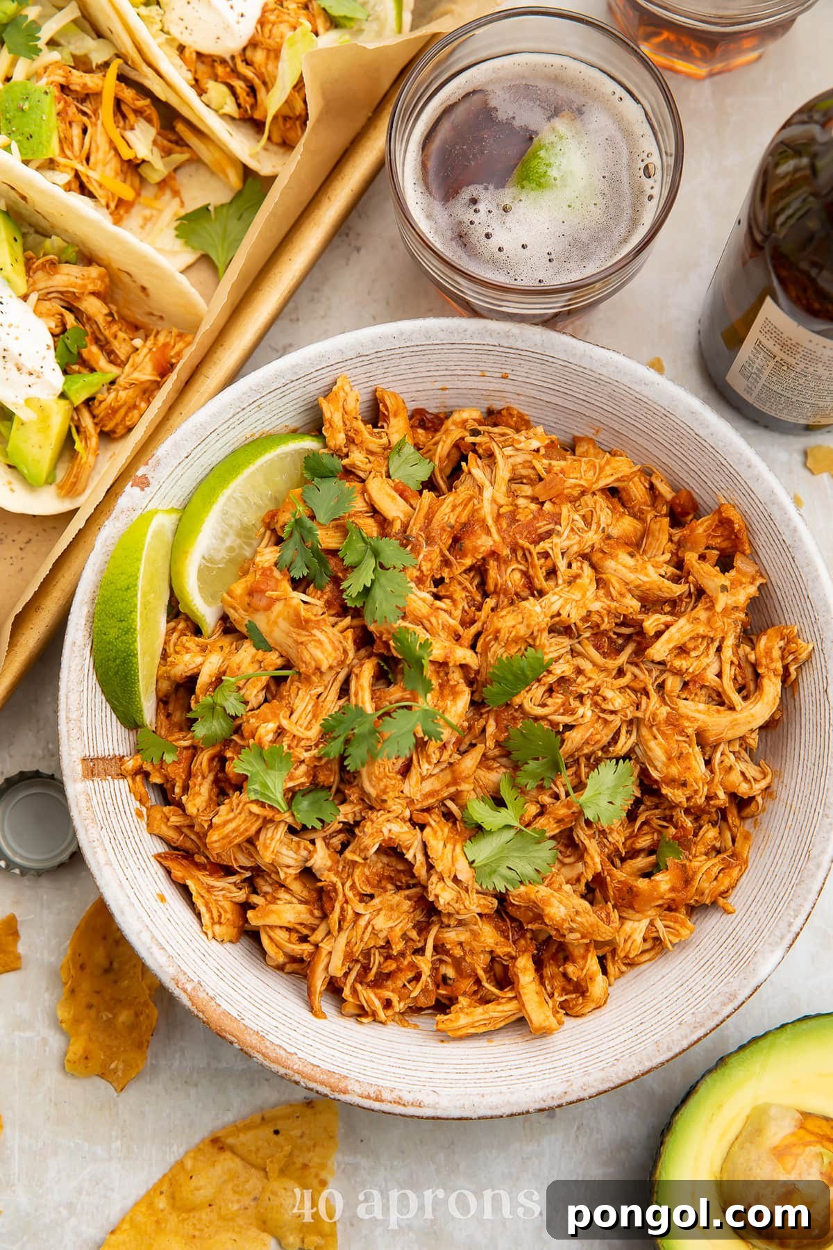 Mexican Chicken Extravaganza 4 Instant Pot salsa chicken, shredded and transferred to a white serving bowl, topped with limes next to a pan of tacos.