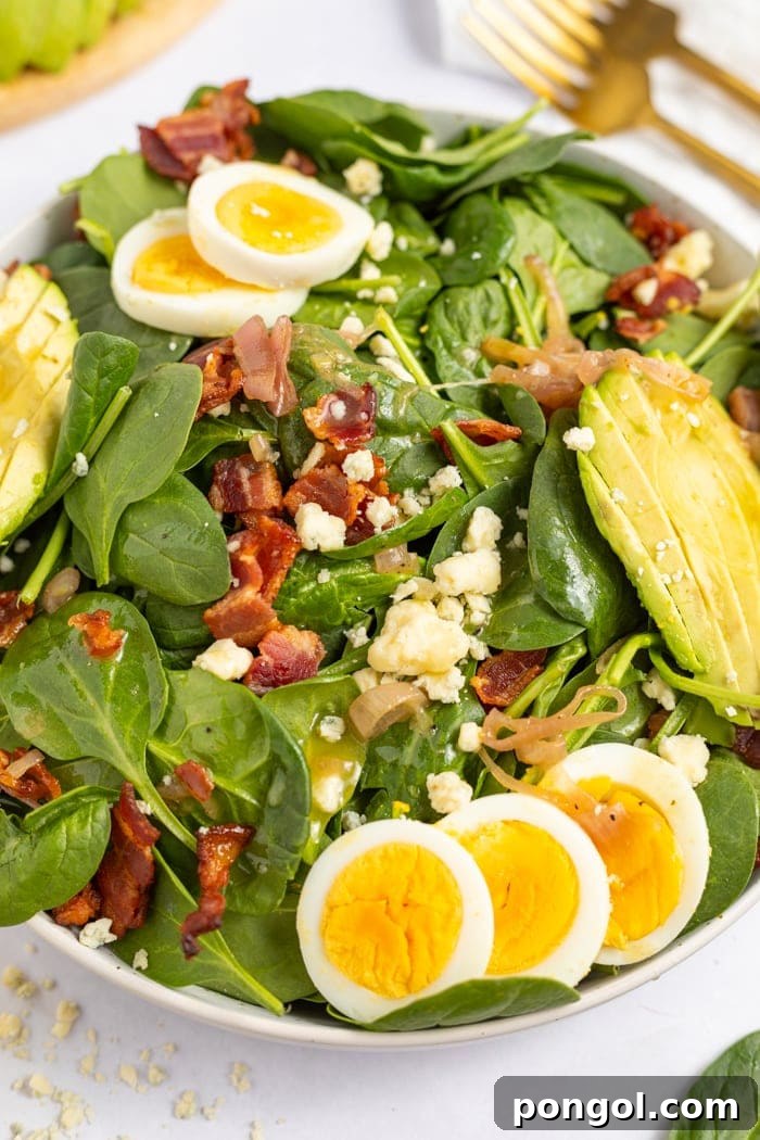 Spinach Salad with Hot Bacon Dressing in a bowl topped with hard boiled eggs, avocado, gorgonzola cheese, and crispy bacon.