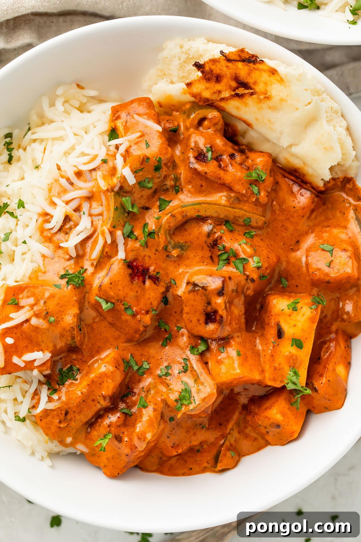 Creamy Paneer Tikka Masala 2 Deep orange paneer tikka masala in a white bowl with white rice and a piece of naan.