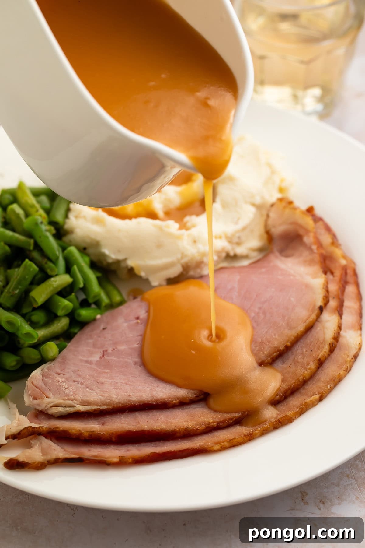 Creamy, richly brown ham gravy being poured from a gravy boat onto slices of ham plated with vibrant green beans and fluffy mashed potatoes.