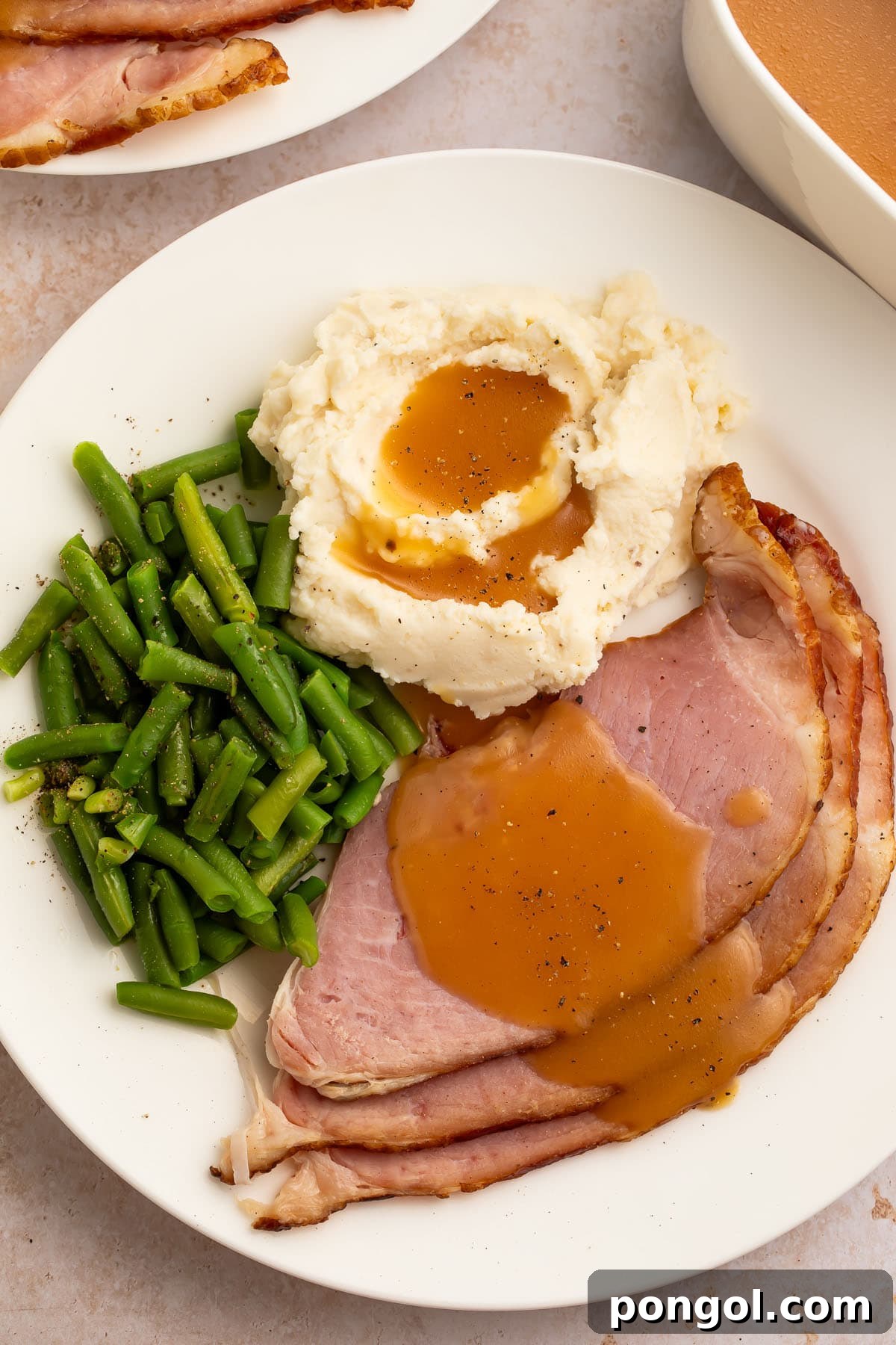 A beautifully plated Easter dinner featuring slices of baked ham generously drizzled with rich, homemade ham gravy, accompanied by vibrant green beans and creamy mashed potatoes.