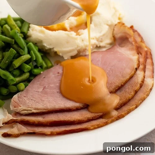 Creamy, richly brown ham gravy being poured from a gravy boat onto slices of ham plated with green beans and mashed potatoes.
