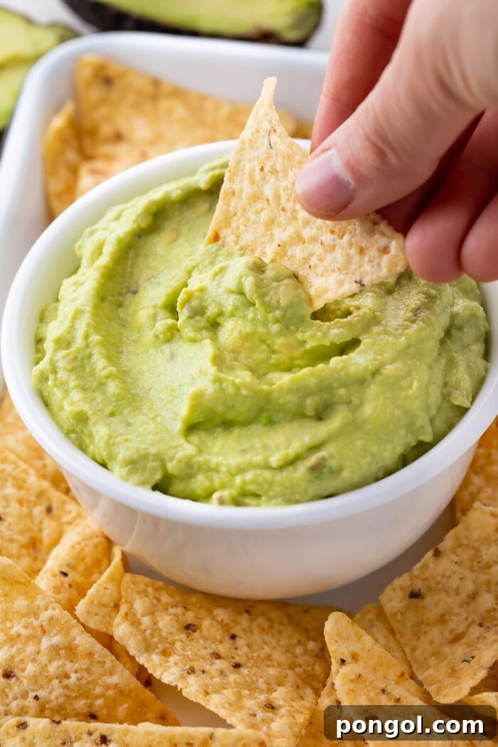 A hand dipping a tortilla chip into a bowl of guacamole dip surrounded by other tortilla chips