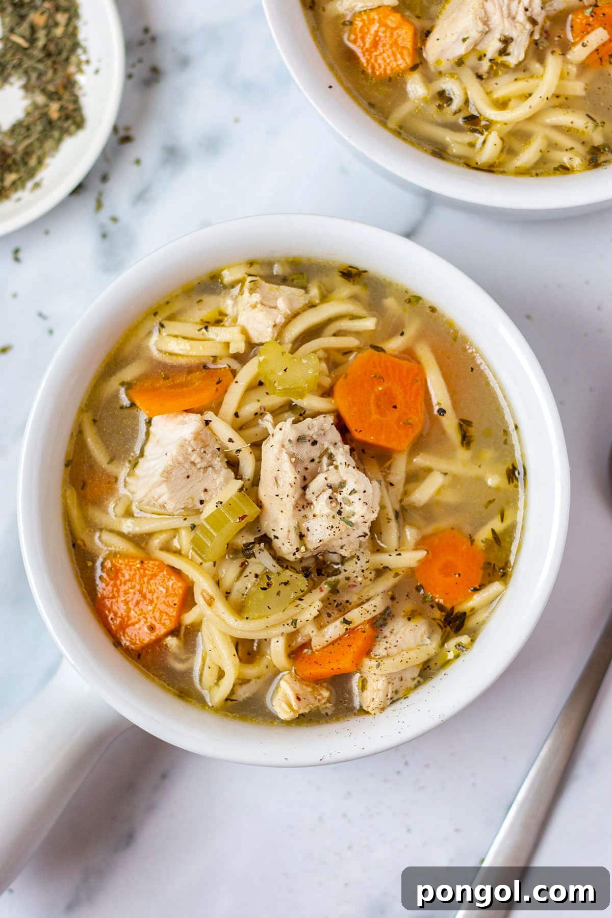 125 Simple Chicken Dinners: Fast Meals with Pantry Staples 126 A small white soup bowl of chunky chicken noodle soup on a white tabletop.