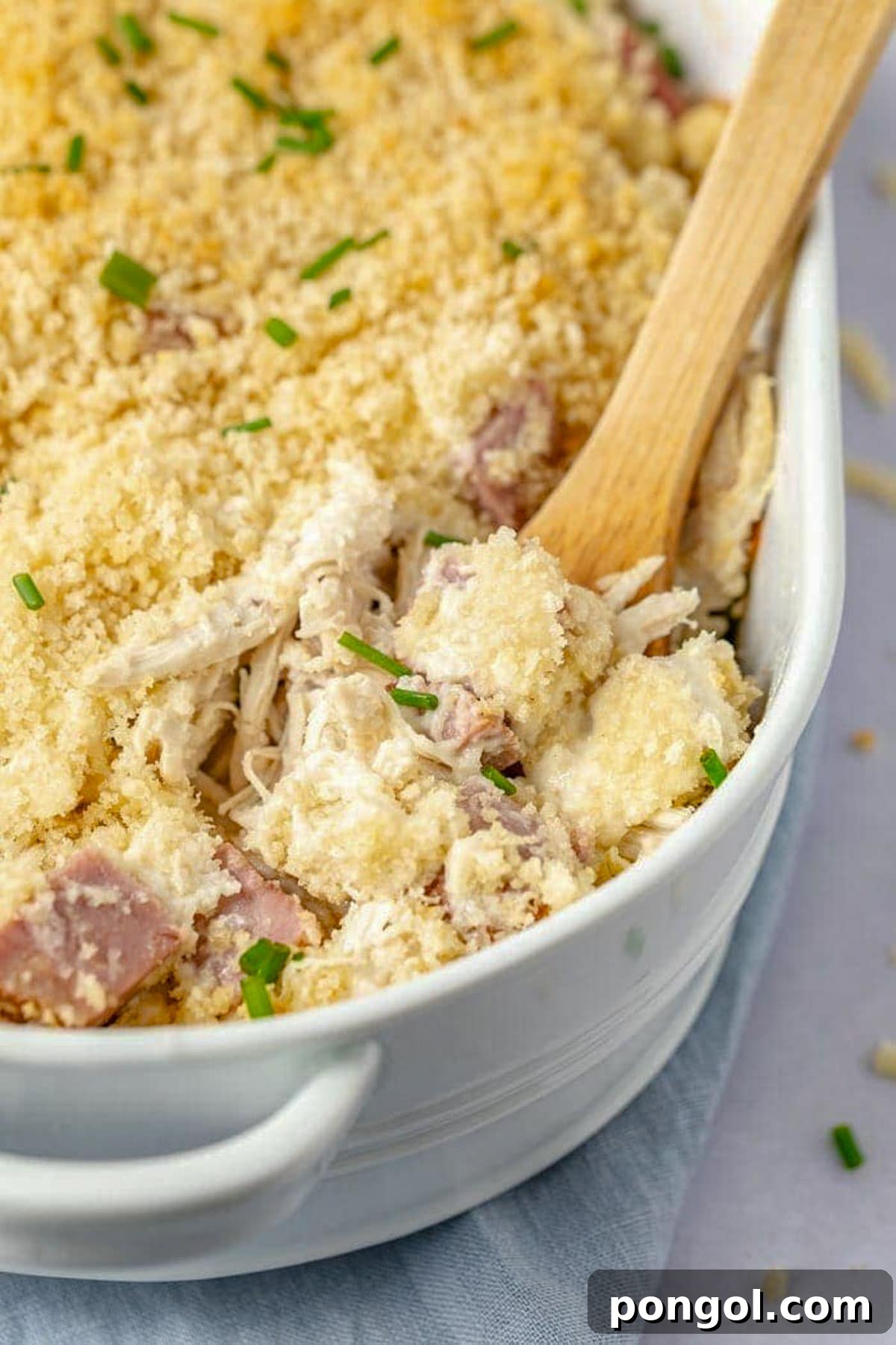 125 Simple Chicken Dinners: Fast Meals with Pantry Staples 19 A chicken cordon bleu casserole in a large white oval casserole dish, with a wooden spoon holding a scoop of casserole resting in the dish.