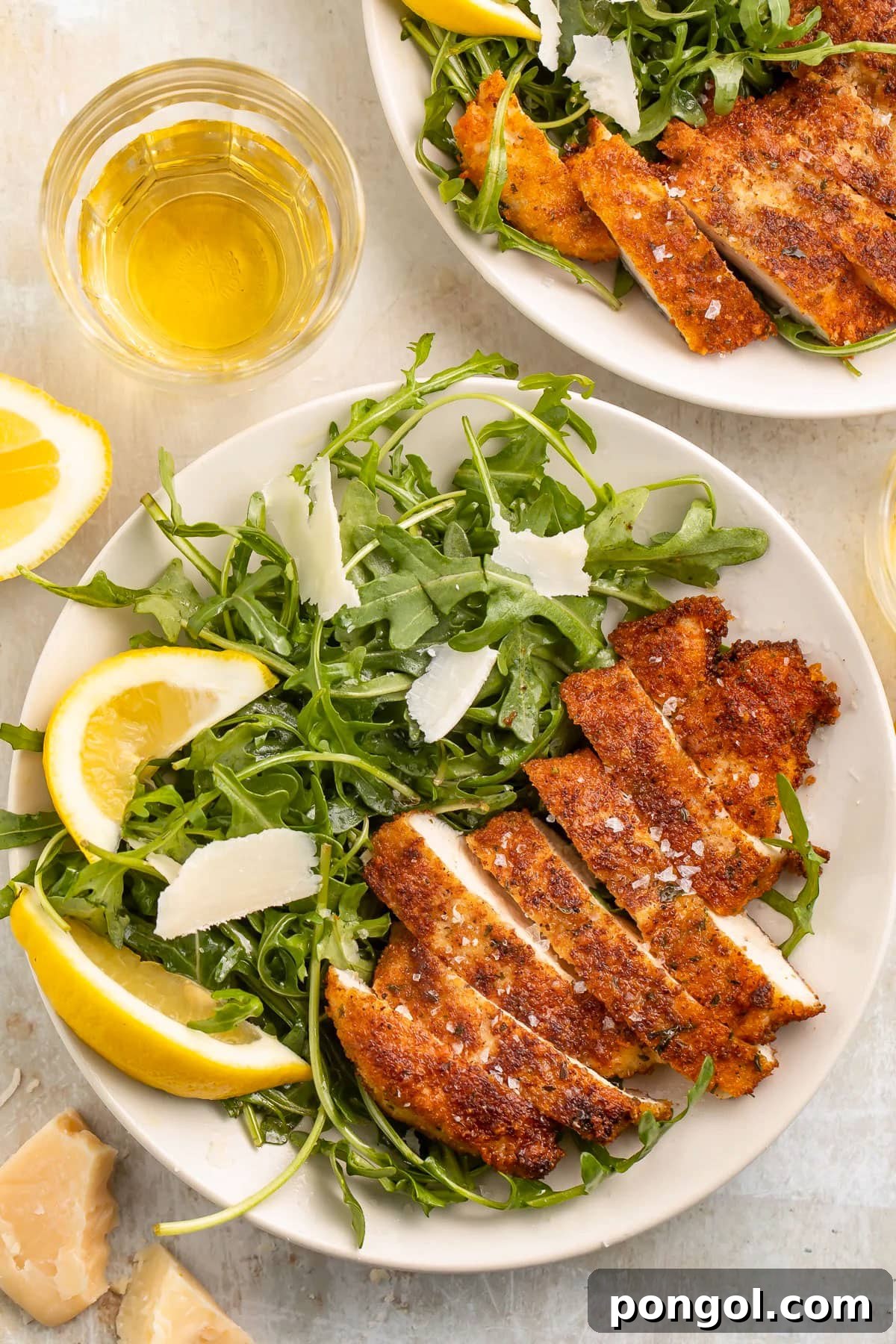 125 Simple Chicken Dinners: Fast Meals with Pantry Staples 21 Top-down view of crispy breaded chicken milanese, sliced and plated next to a small green leaf salad with parmesan shavings and lemon wedges, on a table.