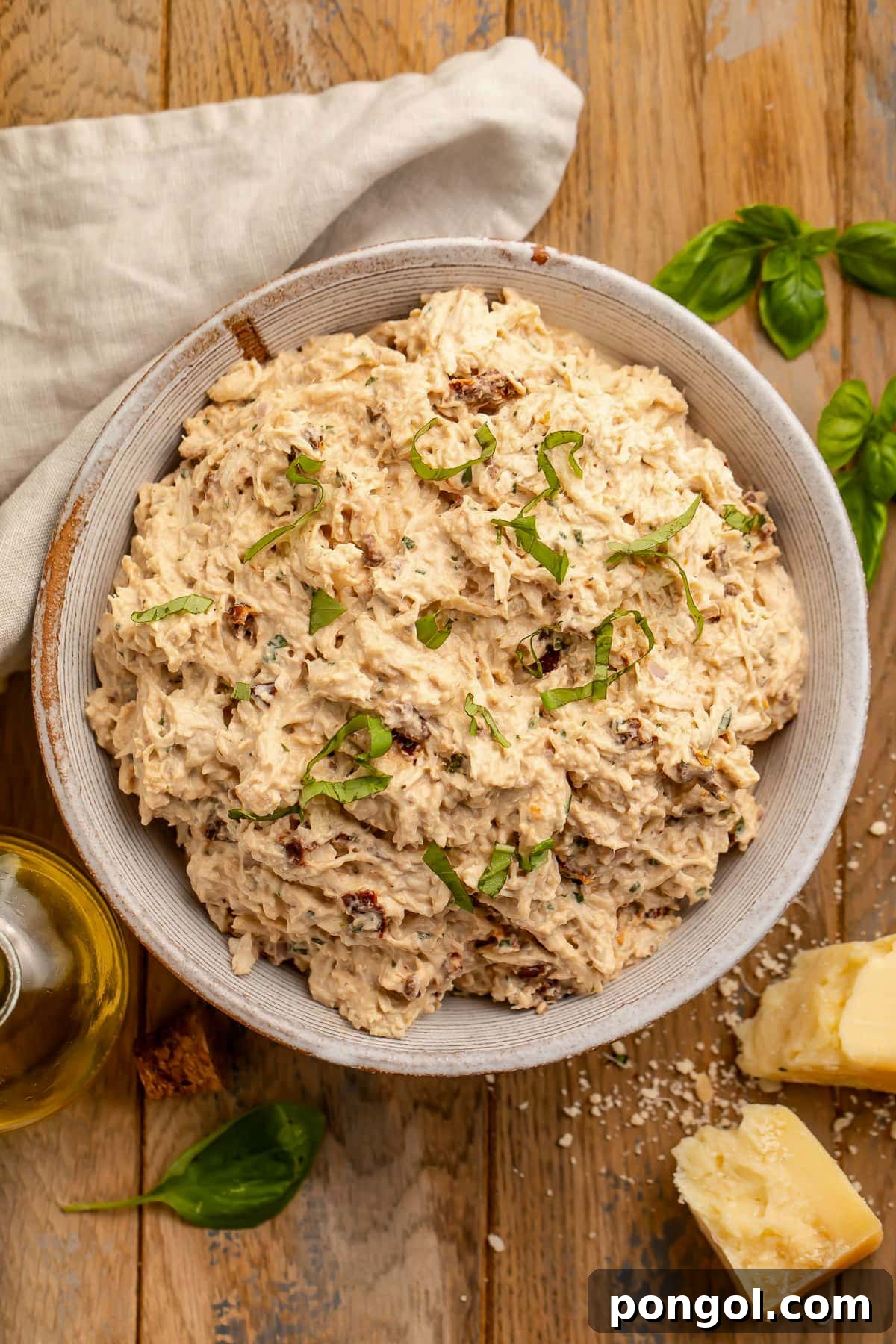 125 Simple Chicken Dinners: Fast Meals with Pantry Staples 45 A large bowl holding creamy marry me chicken salad with shredded chicken, sun-dried tomatoes, and fresh basil on a wooden table.