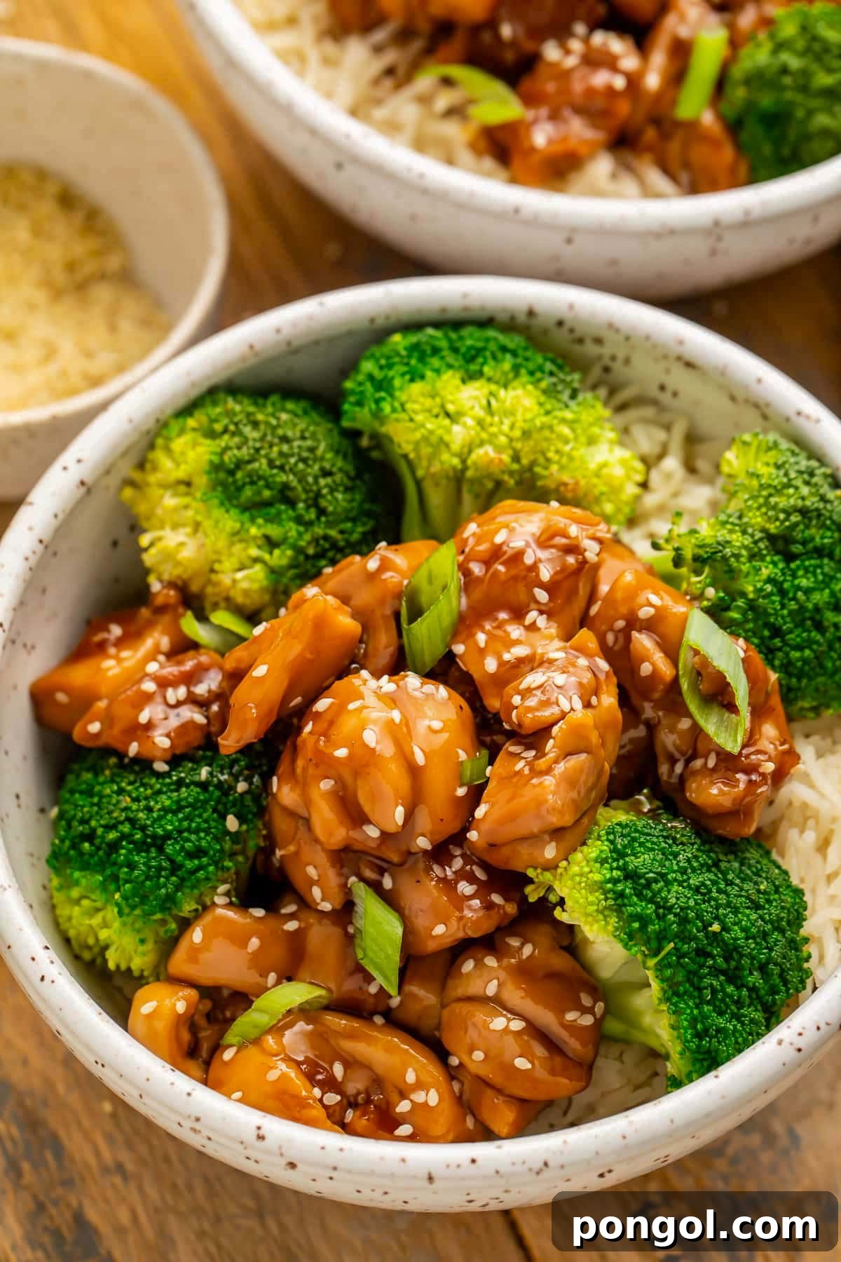 125 Simple Chicken Dinners: Fast Meals with Pantry Staples 62 A white bowl holding white rice, bright green steamed broccoli florets, sticky glazed chicken teriyaki, and a sprinkle of white sesame seeds.