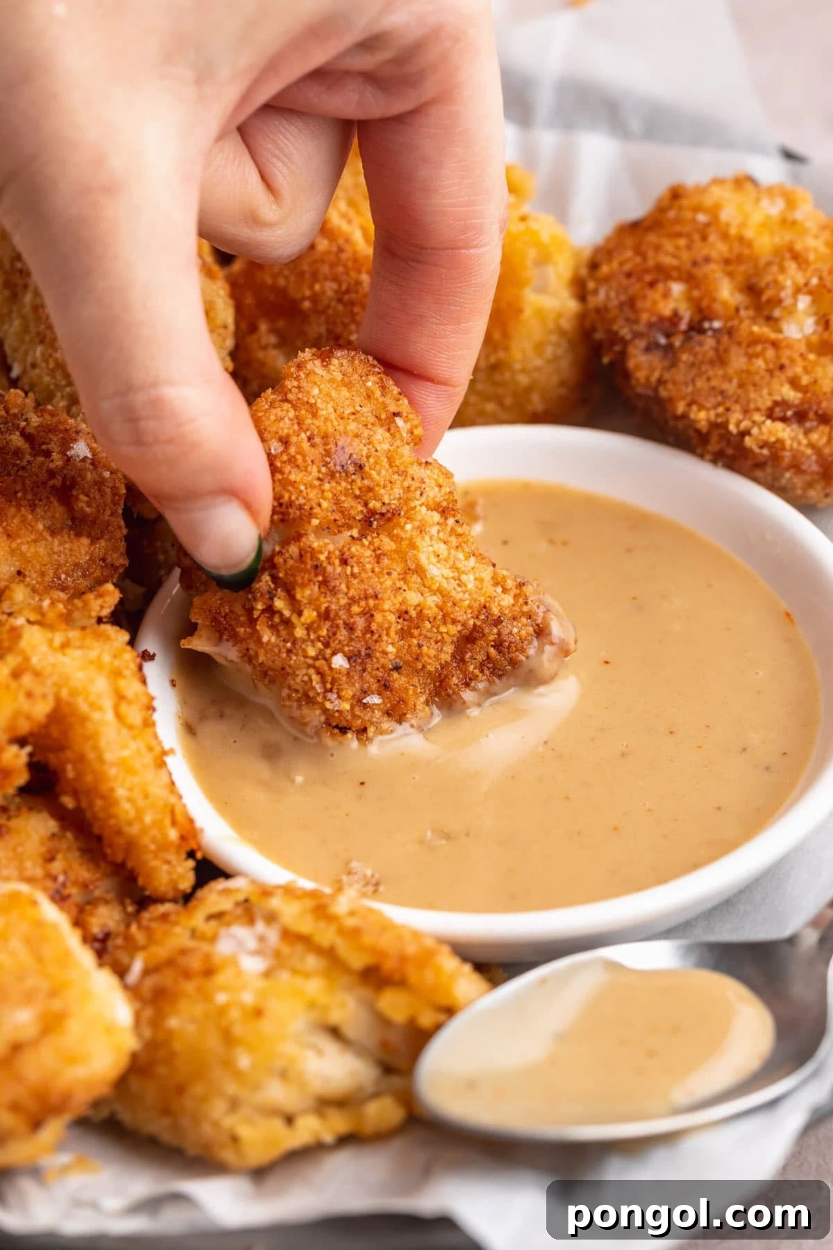 125 Simple Chicken Dinners: Fast Meals with Pantry Staples 76 A white woman's hand dipping a breaded keto chicken chunk into a bowl of keto honey mustard sauce.