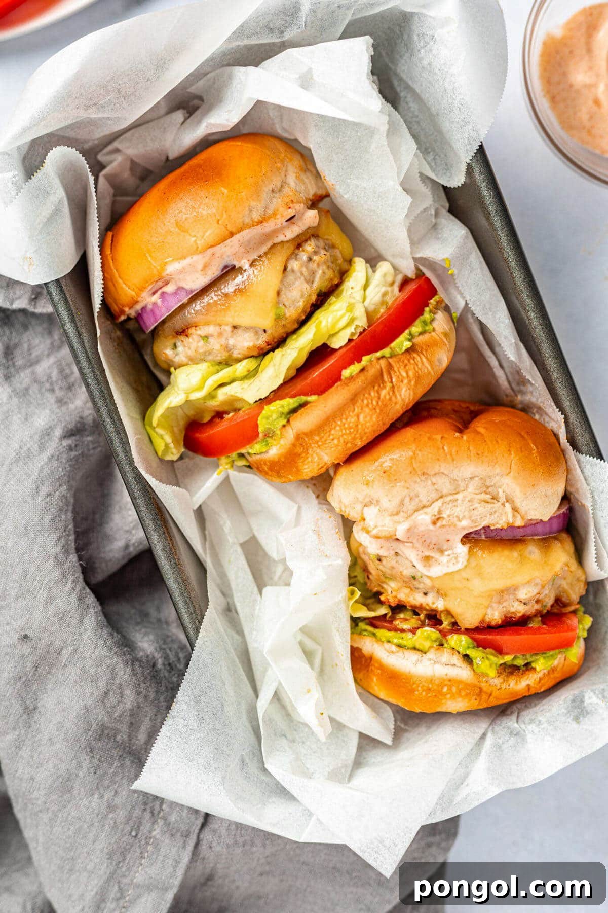 125 Simple Chicken Dinners: Fast Meals with Pantry Staples 99 Two chicken burgers, fully dressed, laying on their sides on a baking pan lined with parchment paper.