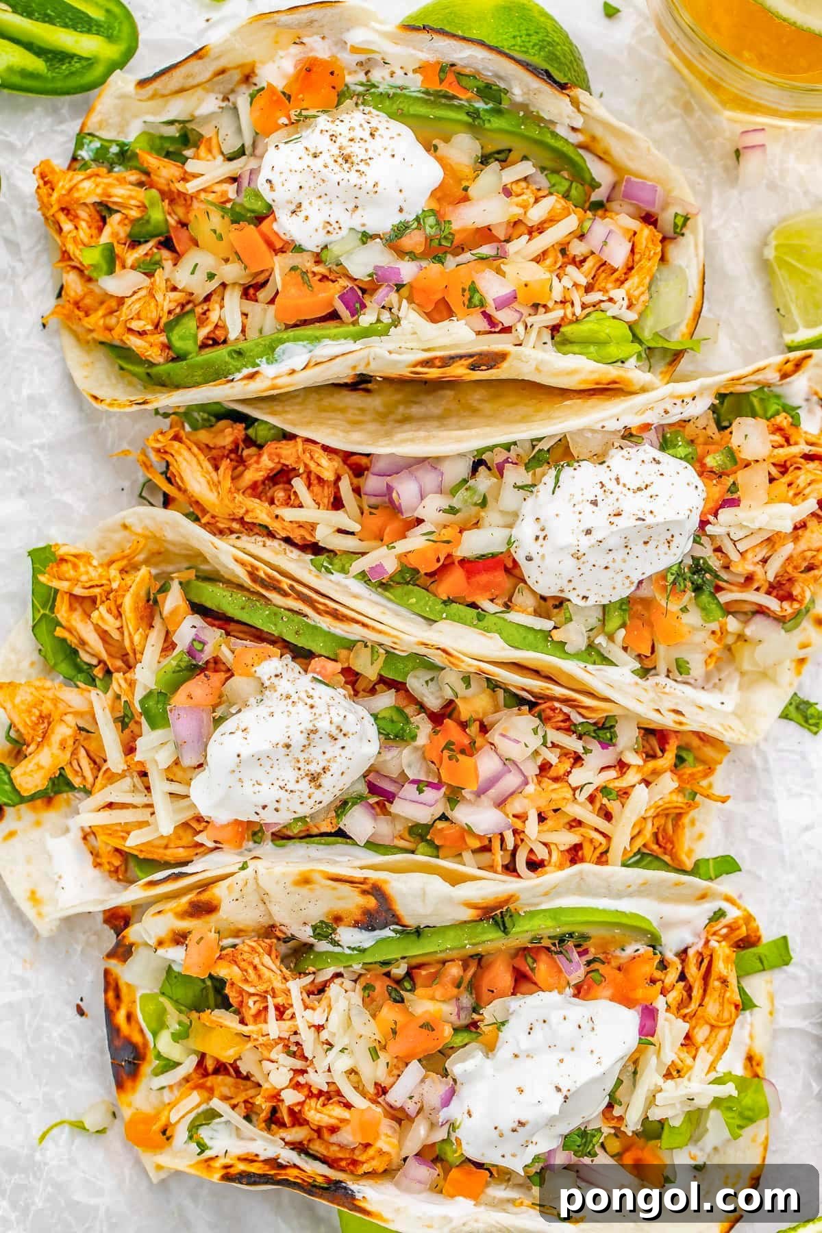 125 Simple Chicken Dinners: Fast Meals with Pantry Staples 100 4 shredded chicken tacos topped with sour cream and toppings, placed side-by-side horizontally in a column on a sheet of parchment paper.