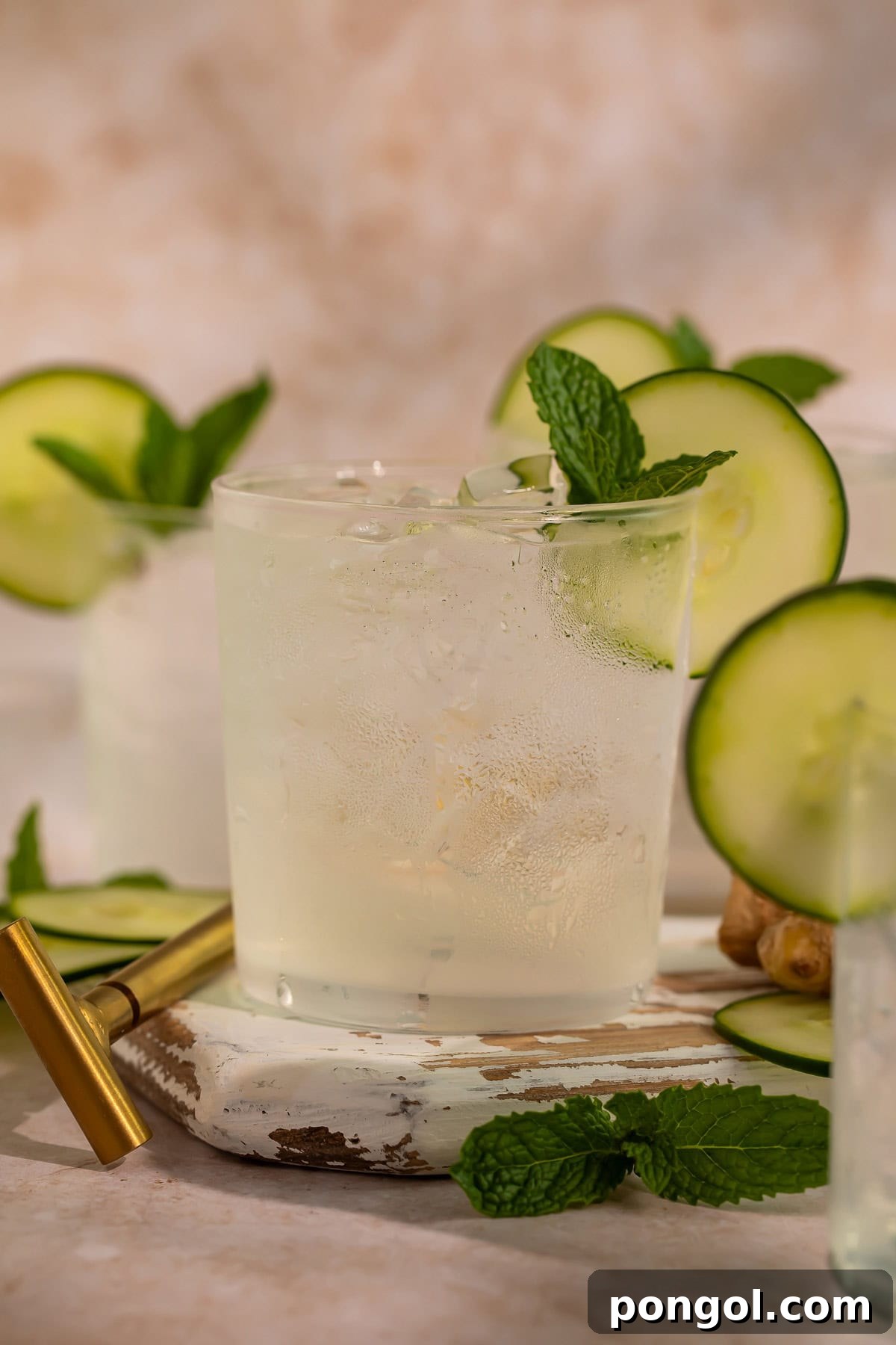 Close-up of a refreshing cucumber ginger mocktail, garnished with cucumber and mint, with another glass in the background, on a light wooden surface.