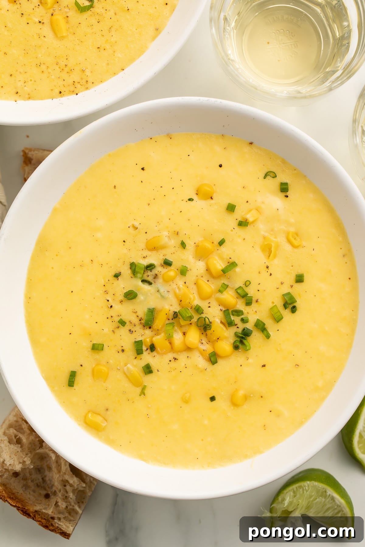 A pristine white soup bowl cradling a generous serving of thick, creamy yellow corn soup. The surface is beautifully garnished with vibrant, whole corn kernels and freshly chopped green onions, inviting a warm, comforting experience.