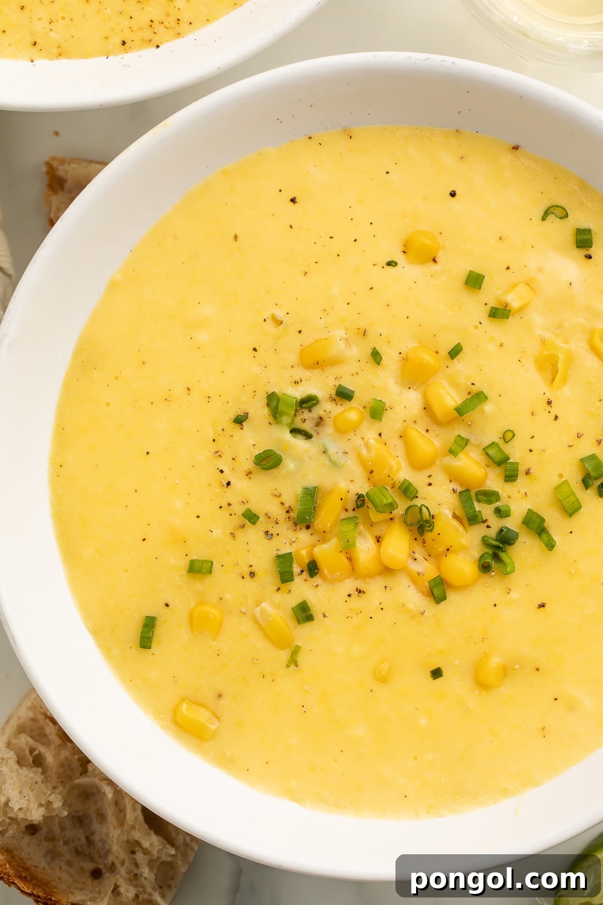 A pristine white soup bowl cradling a generous serving of thick, creamy yellow corn soup. The surface is beautifully garnished with vibrant, whole corn kernels and freshly chopped green onions, inviting a warm, comforting experience.
