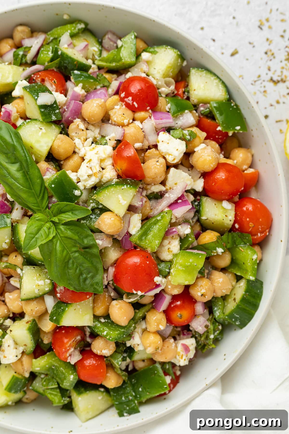 A large white bowl holding a Mediterranean-style chickpea salad with feta, bright red cherry tomatoes, chickpeas, cucumber, onion, and bell pepper.