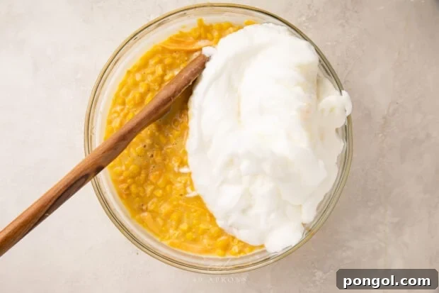Mixture for corn souffle with whipped egg whites in a large glass bowl with a wooden spoon