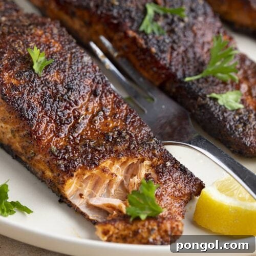 Angled view of blackened salmon on a white plate. The corner of the salmon has been cut into with a fork.