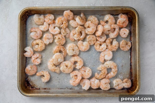 Uncooked shrimp seasoned with olive oil and salt, spread out on a sheet pan, ready for roasting to achieve perfect tenderness.