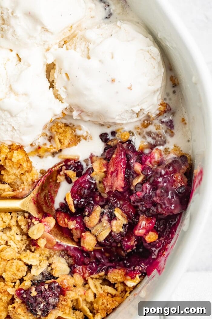 A warm blackberry crumble in a baking dish, ready to be served with a spoon and a side of vanilla ice cream.