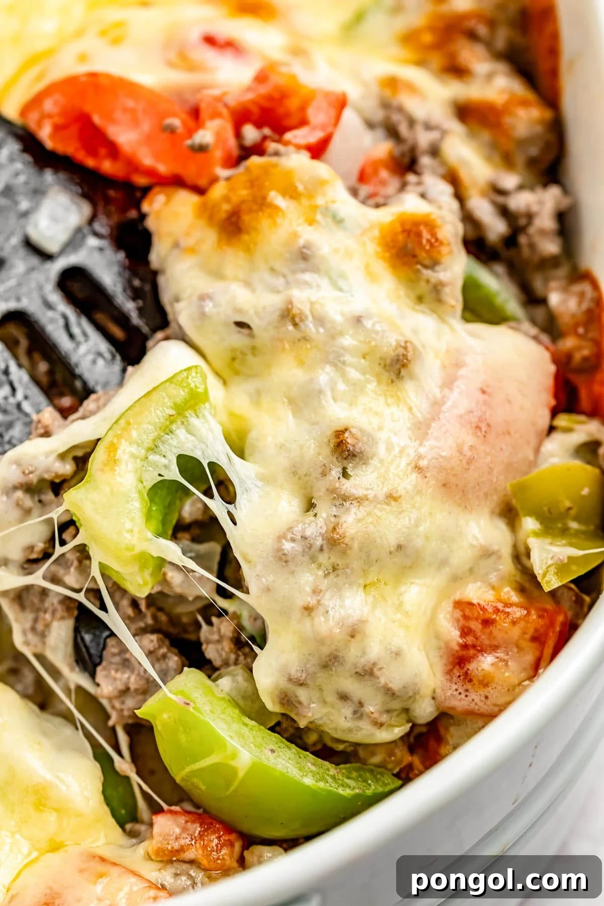 Extreme close-up of a spatula resting in a large casserole dish containing a Philly cheesesteak casseroel with melty cheese, bell peppers, and ground meat.