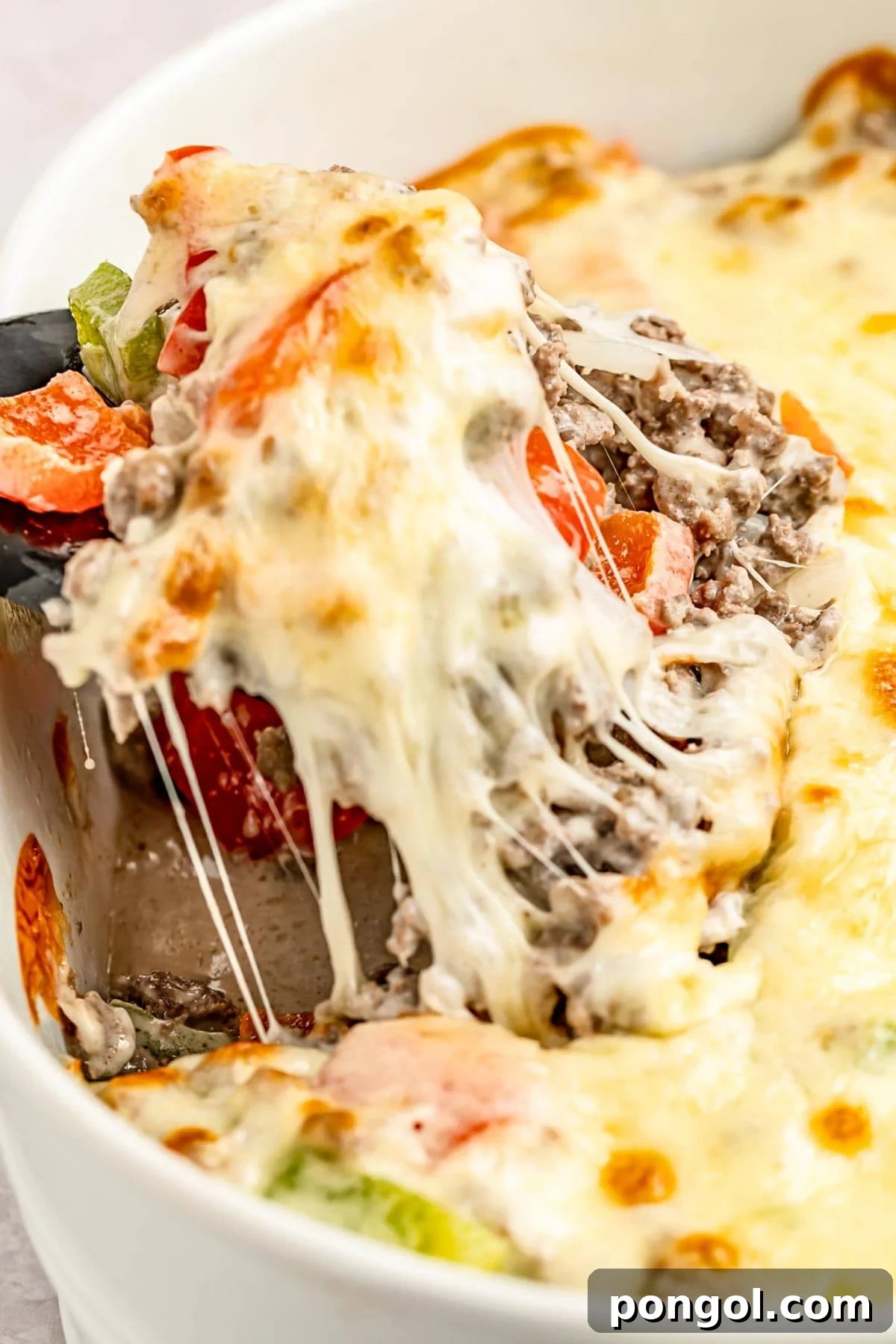 Extreme close-up of a portion of a Philly cheesesteak casserole with melty cheese, bell peppers, and ground meat being lifted out of a casserole dish with a spatula.