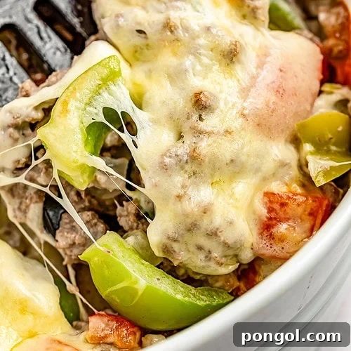 Extreme close-up of a spatula resting in a large casserole dish containing a Philly cheesesteak casseroel with melty cheese, bell peppers, and ground meat.