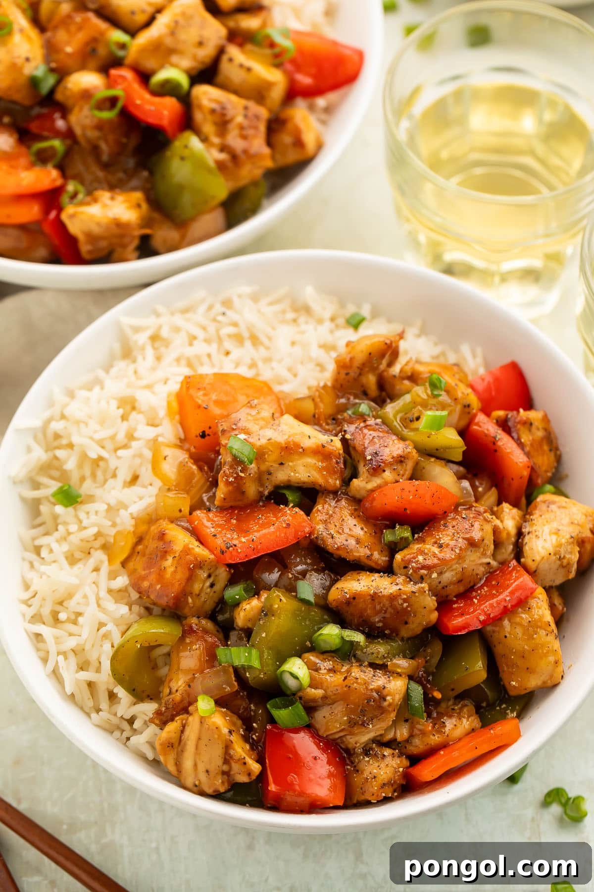Close-up photo of a bowl of white rice topped with black pepper chicken and vegetables on a table, highlighting the glossy sauce and tender pieces.