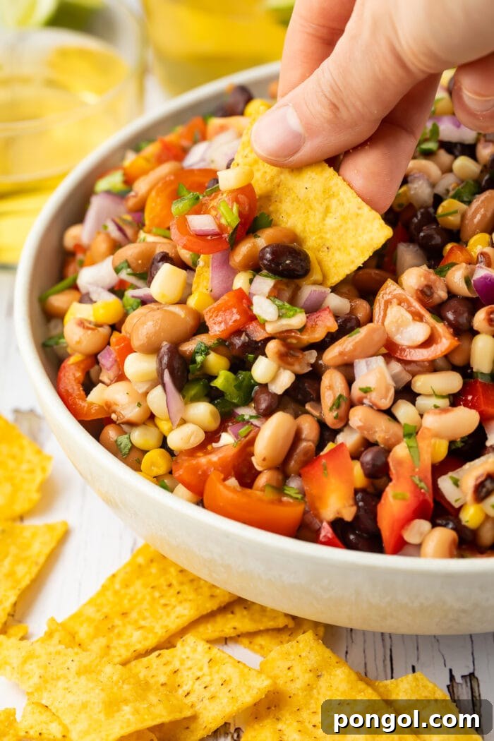 A white woman's hand holds a yellow corn tortilla chip as she uses it to scoop Texas caviar (cowboy caviar) out of a large white bowl.