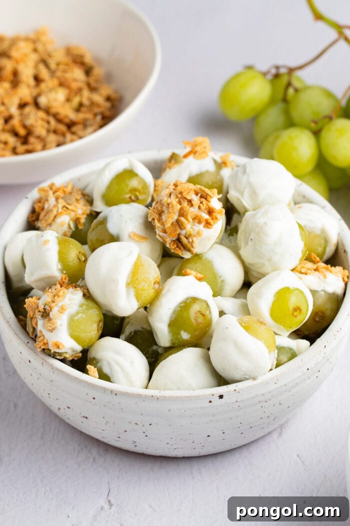 A vibrant display of frozen yogurt-covered grapes in a bowl, with fresh grapes and a separate bowl of granola in the softly blurred background.