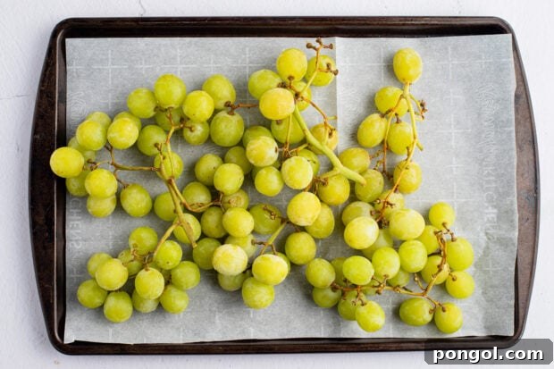 Green grapes neatly arranged on a baking sheet lined with parchment paper, preparing for freezing.