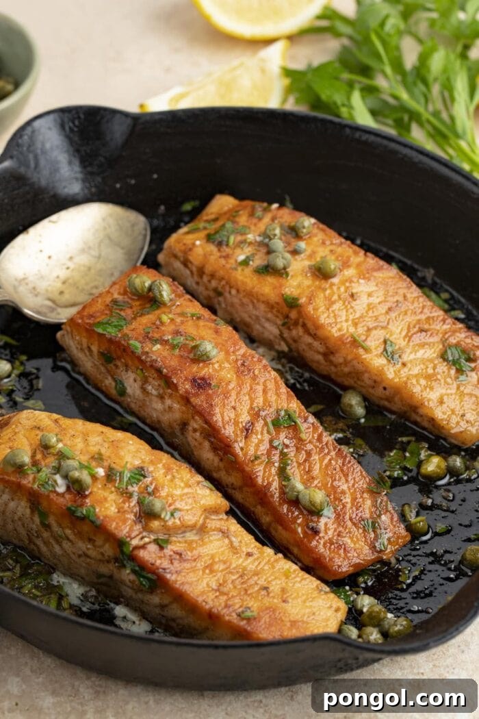 Three perfectly cooked salmon meuniere fillets resting in a cast iron skillet, garnished with vibrant green parsley and bright yellow lemon wedges in the background.