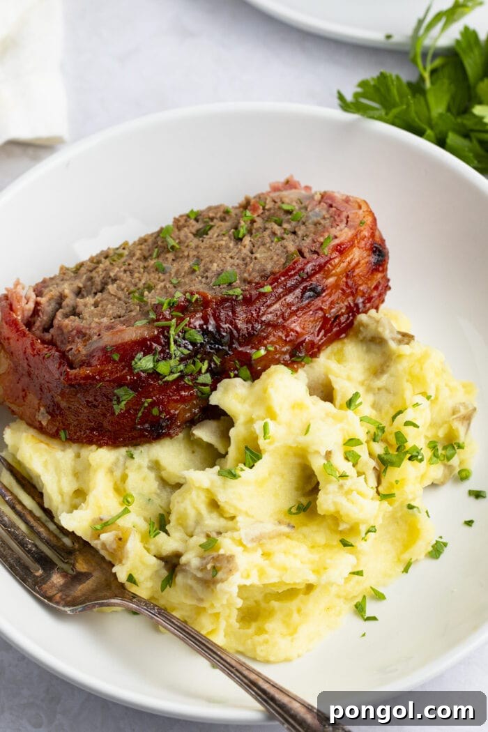 Sliced bacon wrapped meatloaf on a bed of creamy mashed potatoes, garnished with fresh herbs, showcasing the crispy bacon and rich glaze, served on a white plate