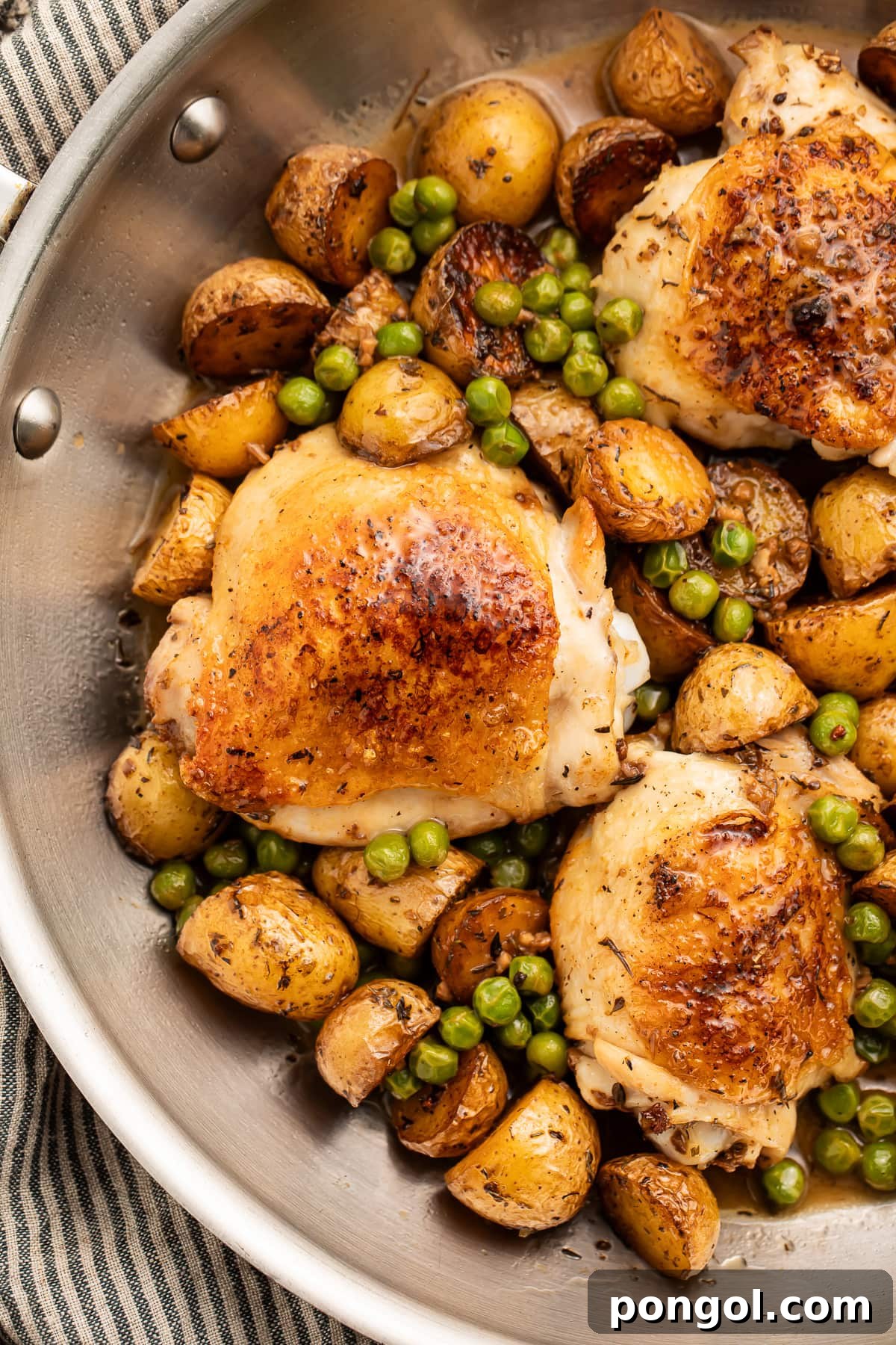 Chicken Vesuvio with golden-brown chicken thighs, roasted potatoes, and bright green peas in a large silver skillet, resting on a striped kitchen towel.