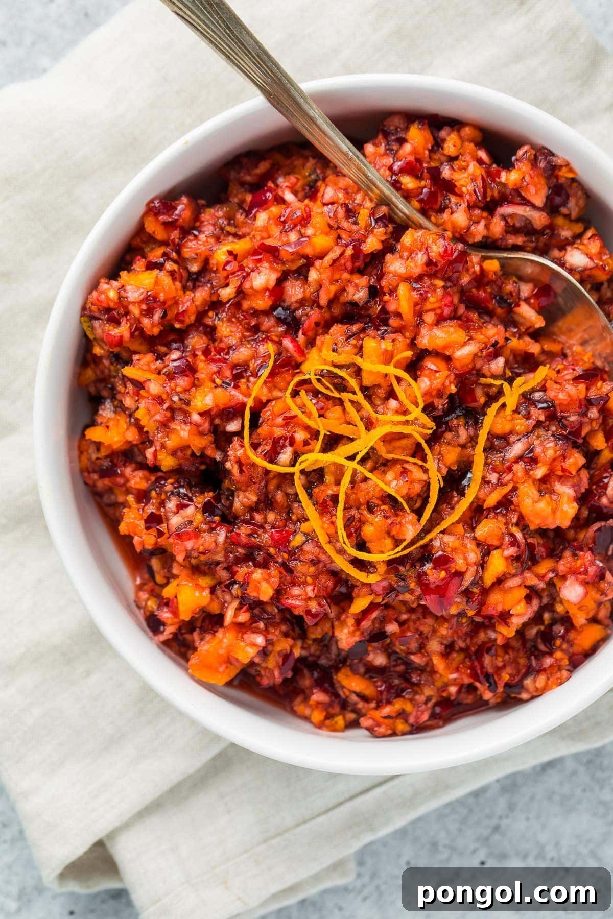 White bowl with vibrant cranberry orange relish, garnished, with a silver spoon on a white napkin.