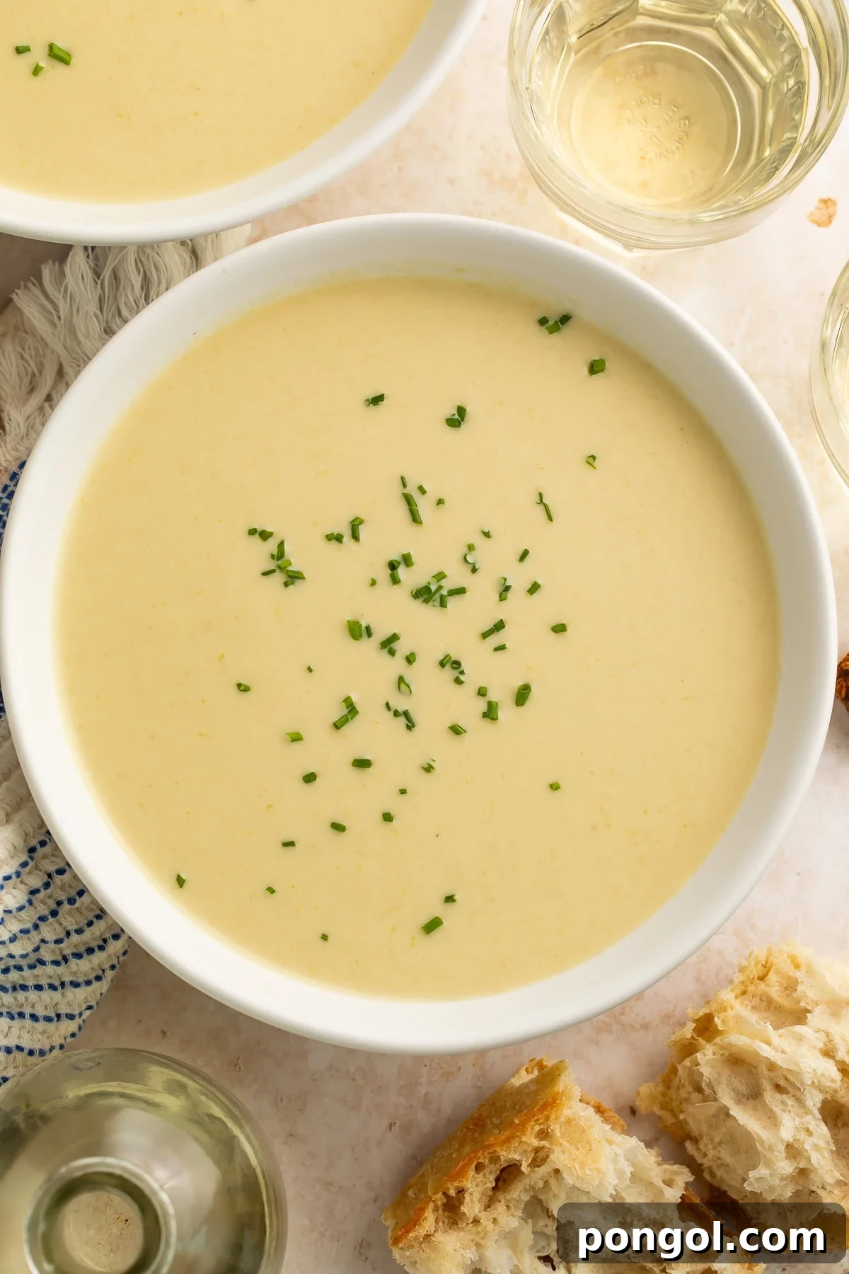 Close-up of creamy celery soup garnished with chives in a white bowl, with bread and wine nearby.