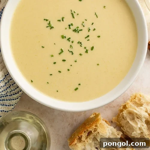 Close-up of creamy celery soup garnished with chives in a white bowl, with bread and wine nearby.